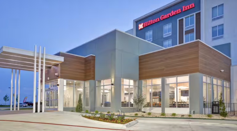 Modern hotel exterior at dusk with large windows and a mix of wood and glass design elements. The Hilton Garden Inn logo is illuminated on the building above. Simple landscaping with small plants is visible in the foreground.